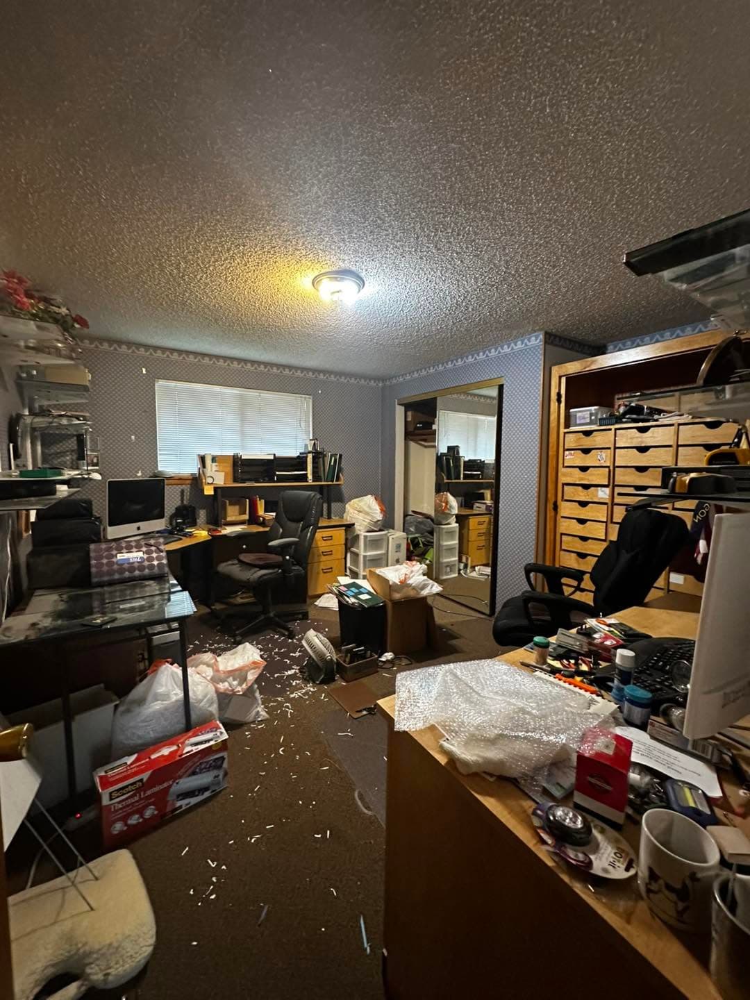 Cluttered home office with multiple desks, scattered items, and shelves of supplies.