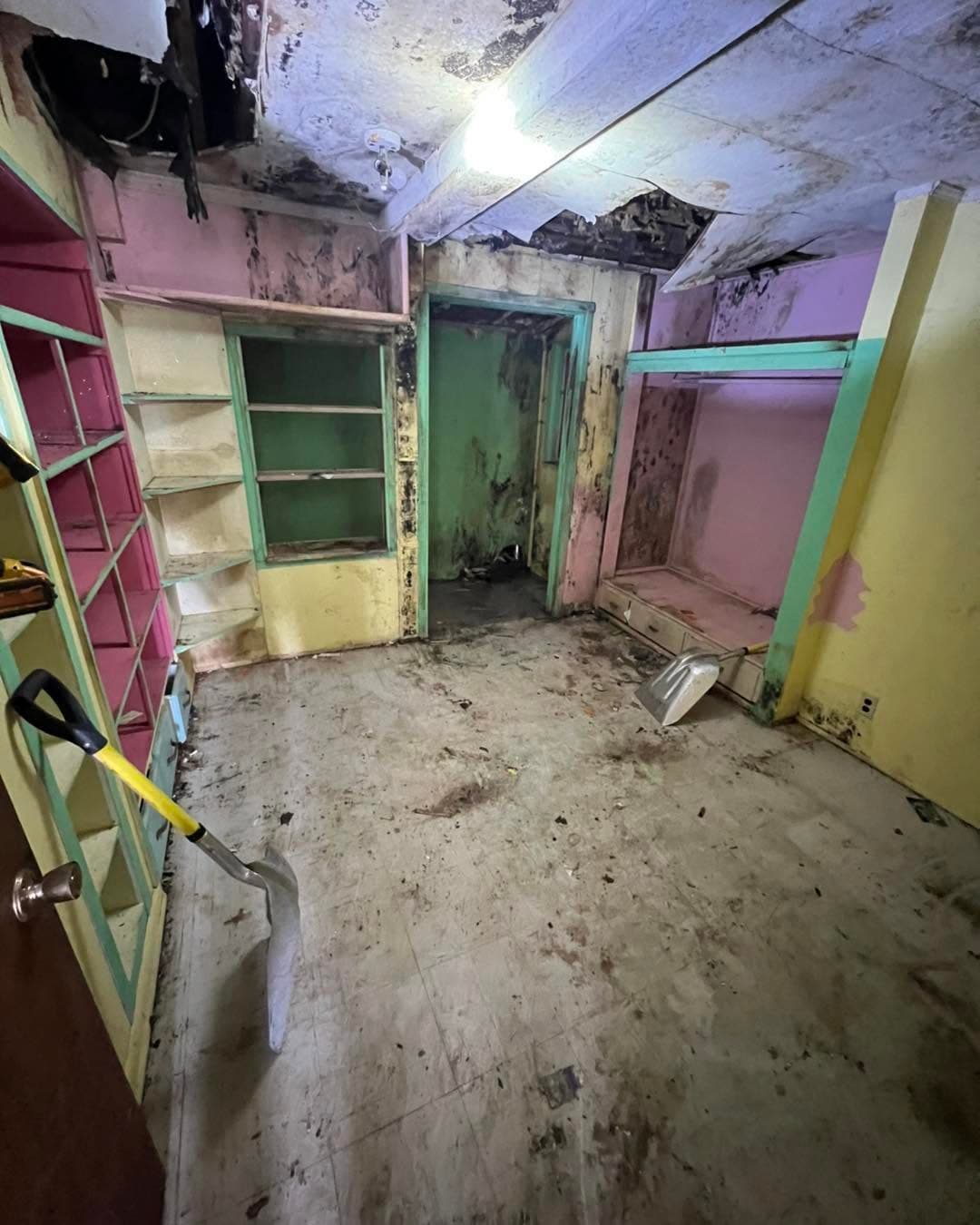 Moldy abandoned room with peeling paint, damaged ceiling, and cluttered floor.