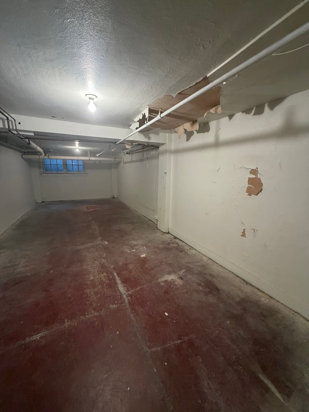 Empty basement with peeling paint, exposed pipes, and a single light bulb overhead.